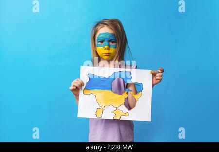 Portrait of a little girl with a painted Ukrainian flag on her face, standing on a blue background and holding a burnt map of Ukraine in her hands. Ch Stock Photo