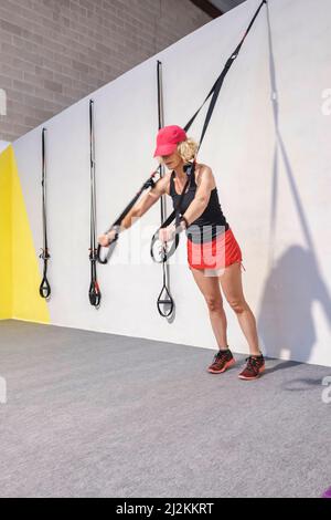 Real young mature caucasian woman in a sport wear in a gym doing push ups training arms with trx fitness straps. Fitness concept. Stock Photo
