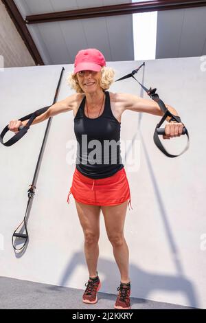 Real young mature caucasian woman in a sport wear in a gym doing push ups training arms with trx fitness straps. Fitness concept. Stock Photo