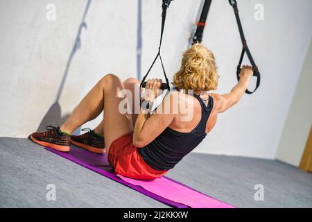 Real young mature caucasian woman in a sport wear at gym training arms exercises with trx fitness straps. Fitness concept. Stock Photo