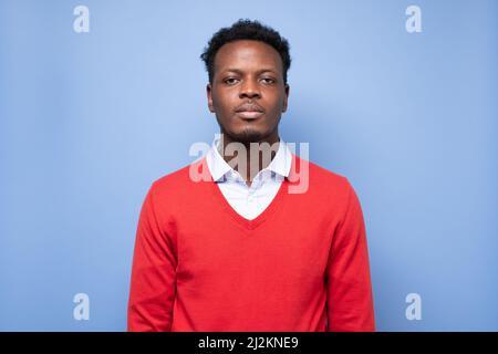 Serious african american millennial man in red sweater isolated on blue studio background posing, Stock Photo