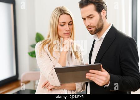 Beautiful woman and serious man with a digital tablet in hands is standing at the office, watching at tablet screen and feeling confused Stock Photo