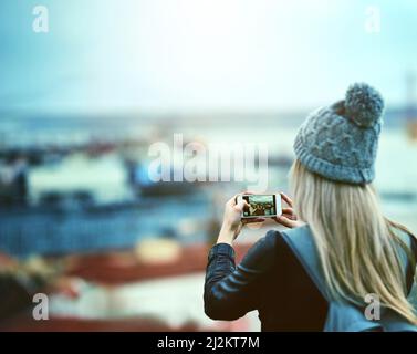 Shes a memory collector. Rearview shot of a young woman using a cellphone to take pictures of a city view. Stock Photo