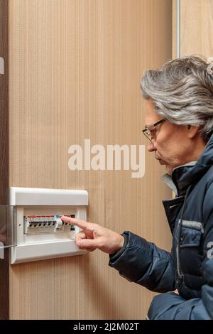 An adult man in a warm jacket looks at the electrical panel. Energy saving concept. Expensive electricity. Stock Photo