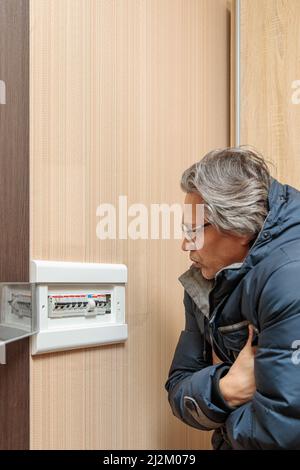 An adult man in a warm jacket looks at the electrical panel. Energy saving concept. Expensive electricity. Stock Photo