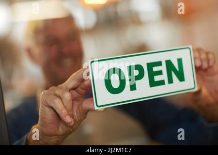 Its official - were open for business. Shot of a happy senior business owner holding up an open sign in the door of his coffee shop. Stock Photo