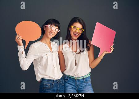 We always think the same. Studio shot of two cheerful young women holding up speech bubbles while standing against a dark background. Stock Photo