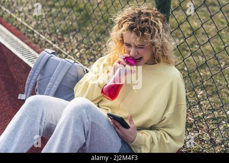 Attractive young Caucasian woman opens the bottle of water on the sports field, holding the phone medium full shot copy space . High quality photo Stock Photo