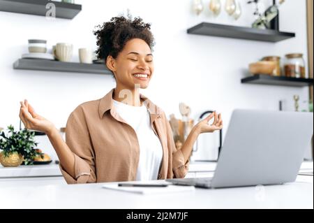 Pretty, positive calm relaxed african american young woman, student, freelancer or designer, working on laptop remotely from home, resting and meditate while work, eyes closed, calmness concept Stock Photo