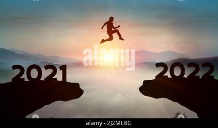 New year concept; pass from 2021 to 2022 , silhouette man jump over barrier cliff and success with beautiful sunrise with cloud background. Starting 2 Stock Photo