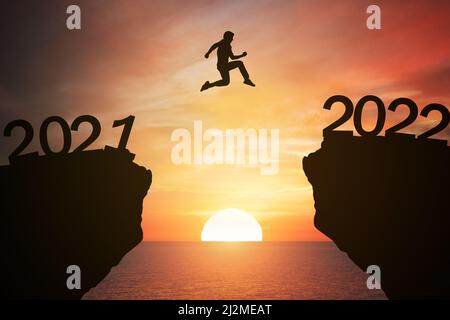 New year concept; pass from 2021 to 2022 , silhouette man jump over barrier cliff and success with beautiful sunrise with sea background. Starting 202 Stock Photo