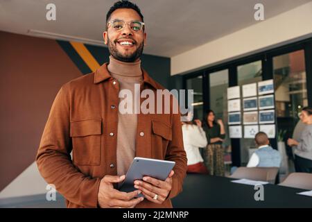 Happy young businessman looking at the camera while holding a digital tablet. Cheerful young businessman standing in a boardroom with his colleagues i Stock Photo