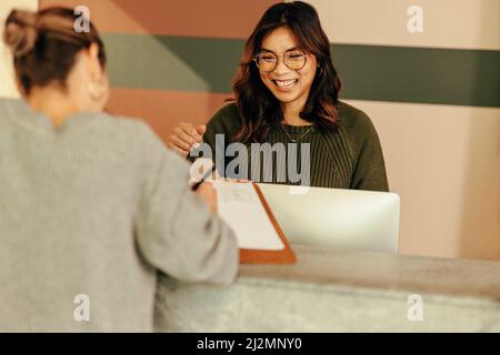 Cheerful receptionist assisting a woman with signing in to an office. Friendly receptionist showing a woman where to sign on a clipboard. Young woman Stock Photo