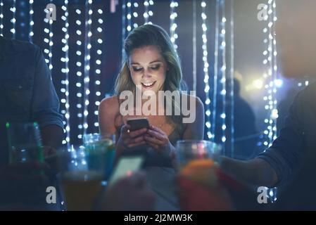 Get here now, this party is awesome. Shot of a young woman texting on her smartphone while sitting in a crowded nightclub. Stock Photo