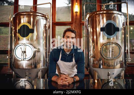 If you want beer we have it. Portrait of a cheerful young barman leaning on the bar counter waiting for customers to serve inside of a beer brewery. Stock Photo
