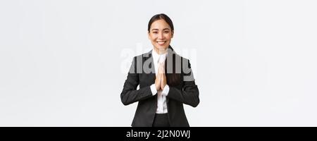 Hopeful glad asian businesswoman in black suit bowing politely and say namaste, greeting clients and smiling friendly. Thankful female manager Stock Photo