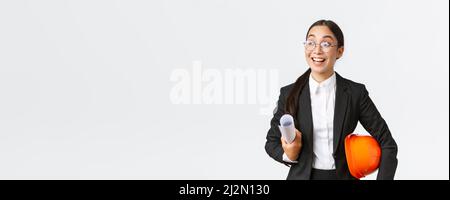 Hopeful and excited asian female architect looking around with fascination and amazement, holding blueprints and helmet, walking in construction area Stock Photo