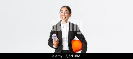 Hopeful and excited asian female architect looking around with fascination and amazement, holding blueprints and helmet, walking in construction area Stock Photo