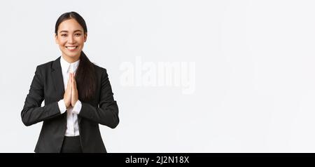 Hopeful glad asian businesswoman in black suit bowing politely and say namaste, greeting clients and smiling friendly. Thankful female manager Stock Photo