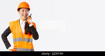 Confident pleased asian female engineer in safety helmet and uniform talking with chief architect using walkie-talkie. Satisfied construction Stock Photo