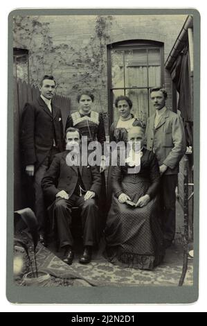 Original vintage 1900s photo Victorian / Edwardian portrait middle class Victorian family group outside in their small garden/yard. 2 married couples - wives with dapper husbands, maybe brothers, mother & father in law sitting at the front. The men are smartly dressed with suits, ties, matching collars, moustaches. The older couple look as if they are from the Victorian era, but the younger people wear more fashionable early Edwardian attire - several generations, multigenerational Victorian family, Victorian mother, father, Victorian parents. 1900's husbands & wives. Circa 1901, 1902, 1903 UK Stock Photo