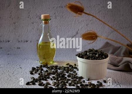 Sunflower seeds in a white ceramic bowl and in bulk on a table with a glass bottle of sunflower oil on a gray background with dry plants Stock Photo