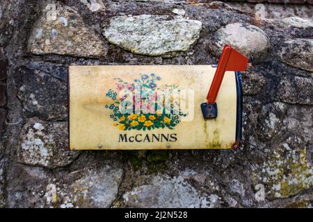 Old weathered metal mailbox mounted on gray stone wall with red flag and flowers paint motive. Ireland Stock Photo