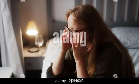 Young caucasian woman with long, ginger head sitting on her bed, rubbing her temples, because of another migraine headache . High quality photo Stock Photo