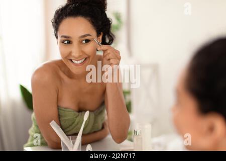 Smiling young woman wearing towel, correcting eyebrows with pincers near mirror at home, free space Stock Photo