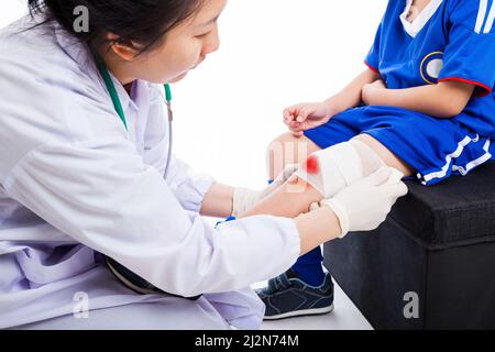 Youth asian (thai) sport boy in blue uniform. Doctor perform first aid knee injure by bandage, knee have bruise and bloody on bandage (knee  in focus) Stock Photo