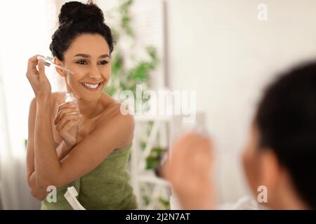 Attractive young brunette woman applying moisturizing serum onto her face near mirror at home, free space Stock Photo