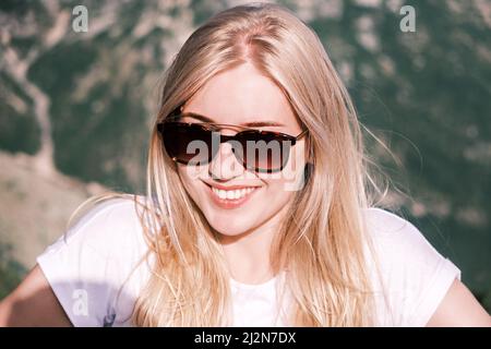 Portrait confident young cheerful smiling blonde woman looks into the camera wearing sunglasses against the background of nature on a sunny day. Happy Stock Photo