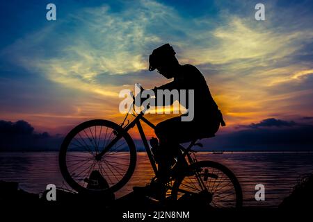 Silhouette of bicyclist riding the bike on a rocky trail at seaside, on colorful sunset sky background. Active outdoors lifestyle for healthy concept. Stock Photo