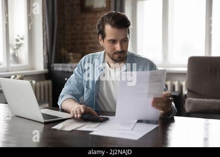 Focused millennial man paying domestic bills, using calculator Stock Photo