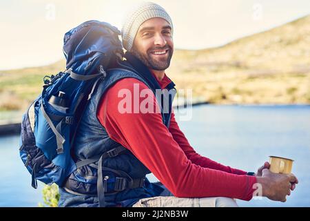 All I need is coffee and a view. Portrait of a happy hiker drinking coffee while admiring a lake view on a trail. Stock Photo