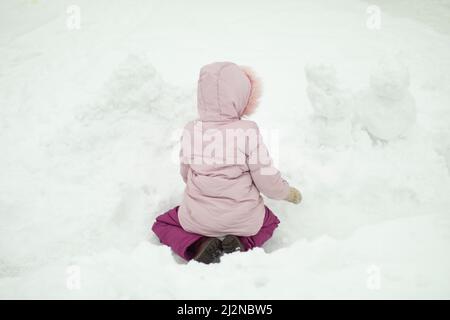 Child sculpts snowman. Girl plays in snow. Preschooler plays outside in winter. Warm clothes. Stock Photo