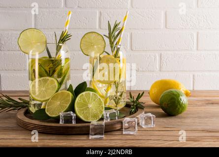 two glasses of refreshing citrus water, a cocktail or tonic with ice cubes on a wooden table against a white brick wall. useful detox Stock Photo