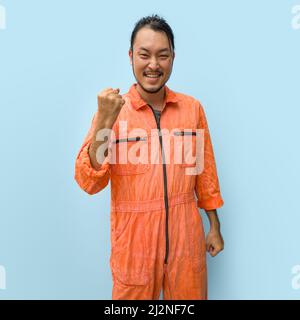 Young long hair asian man with mustache and beard stand smiling while raise the fist up in front. Portrait of industrial people on blue background. Stock Photo