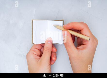 Crop view of female hands making notes with a pencil in a miniature book with blank pages against a concrete table background. Creative process concep Stock Photo