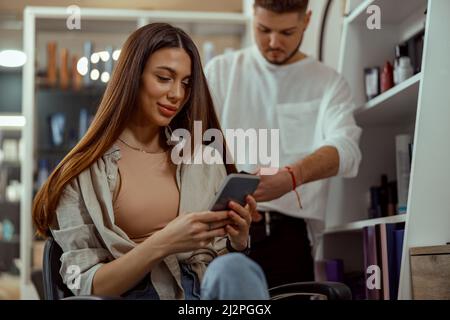 Happy woman using smart phone while getting her hair styled at beauty salon Stock Photo