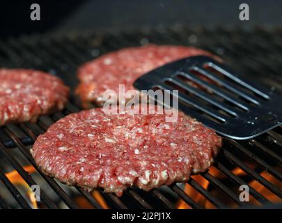 Fresh raw beef burgers on grill grate with flames and stainless steel spatula in background Stock Photo
