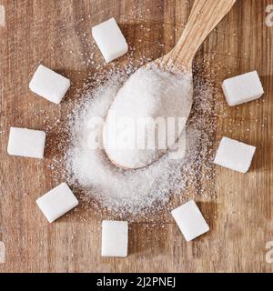 The wooden spoon full of granulated sugar and sugar cubes around on wooden background. Stock Photo