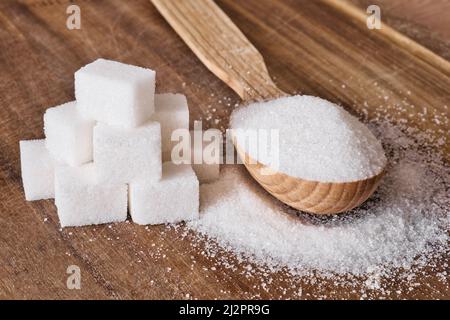 Granulated sugar in wooden spoon and sugar cubes stacked in pyramid on wooden background. Stock Photo