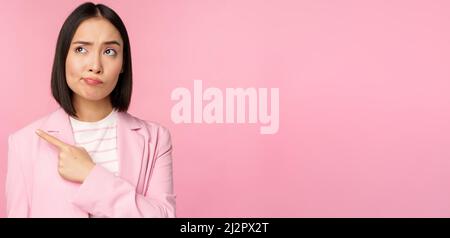Doubtful business woman, sulking and grimacing while pointing, looking left at banner with skeptical face expression, posing against pink background Stock Photo