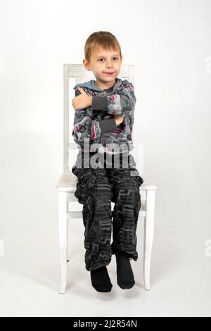 The boy sits with his arms around himself on a chair on a white background. Stock Photo