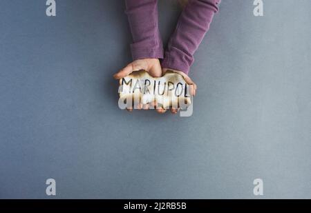 children's hands hold a burnt piece of paper with the inscription Mariupol. the war in Ukraine burned down the city. Stock Photo