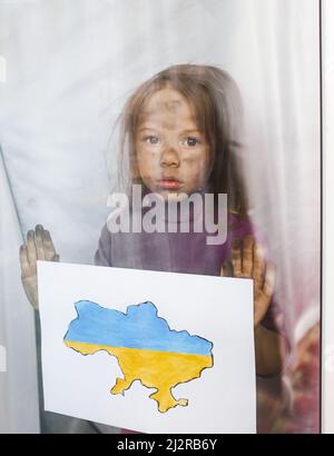 a little Ukrainian girl with sad eyes stands behind the glass, holding a drawn map of Ukraine in her hands. Children and war. Stock Photo