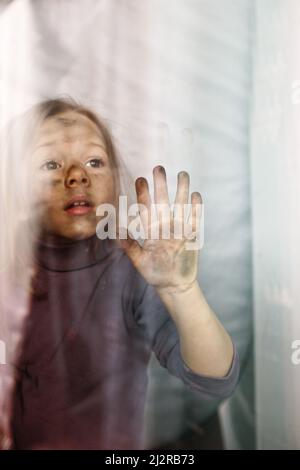 a little Ukrainian girl with a face blackened by fire is standing behind a glass with sad eyes. Children and war. Stock Photo