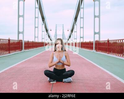 beautiful, sporty, fit, young caucasian woman in sportswear and sneakers sitting in lotus pose on the bridge. Sport, activity, yoga, urban lifestyle c Stock Photo