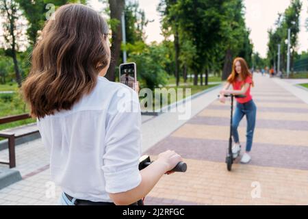 Two girl friends riding push e scooter and do selfie via their smartphones outdoors. Group gen z young people using electric scooter in city park Stock Photo